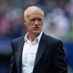 France national team manager Didier Deschamps with short white hair wearing navy suit jacket and white open collar shirt Close up shot on football pitch sideline with blurred stadium crowd and blue seating in background Deschamps has focused expression during match  trybalnews
