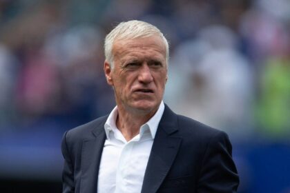France national team manager Didier Deschamps with short white hair wearing navy suit jacket and white open-collar shirt. Close-up shot on football pitch sideline with blurred stadium crowd and blue seating in background. Deschamps has focused expression during match.