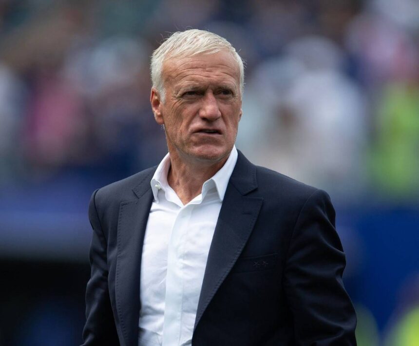 France national team manager Didier Deschamps with short white hair wearing navy suit jacket and white open collar shirt Close up shot on football pitch sideline with blurred stadium crowd and blue seating in background Deschamps has focused expression during match  trybalnews