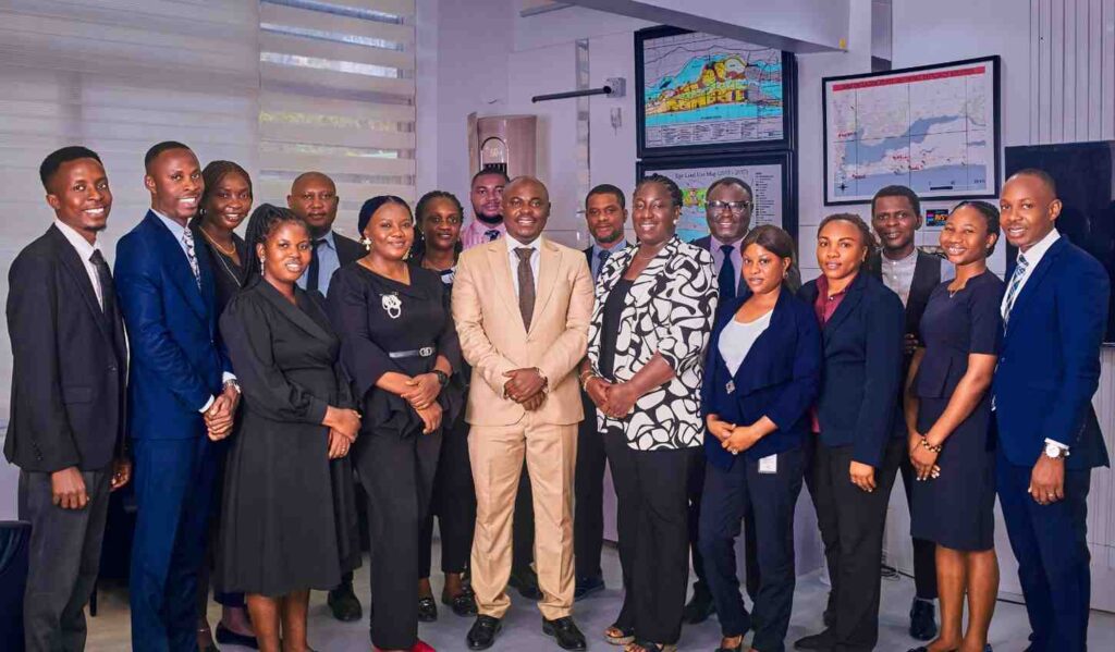 EFFICACYDEVELOPMENTPLCSTAFFS Trybal News Managing Director Engr Oyeleke Ajiboye in a tan suit and brown tie stands at the center of a group of 16 Efficacy Development Plc staff members The team men and women in business formal attire including black navy and blue suits and dresses smile for a group photo in a modern office Two framed maps are displayed on the white paneled wall behind them trybalnews