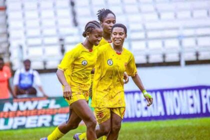Three Edo Queen players in yellow uniforms celebrate together on a grassy field with empty stadium seats in the background.