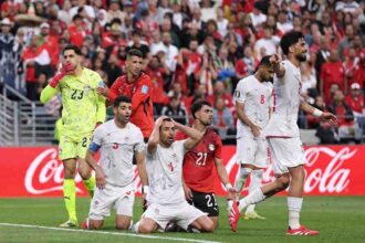 IR Iran players in white kits with red numbers show frustration after a missed chance. Player #4 kneels on the grass with both hands on his head, while teammate #5 kneels beside him with a shocked expression. Teammate #11 stands to the right pointing and shouting. Egypt goalkeeper #23 in a neon yellow kit and defender #21 in red watch on. 2026 FIFA World Cup Group G match between Egypt and IR Iran on June 26, 2026 at Seattle Stadium in Seattle, Washington. A red Coca-Cola hoarding and packed crowd in red and white are in the background.