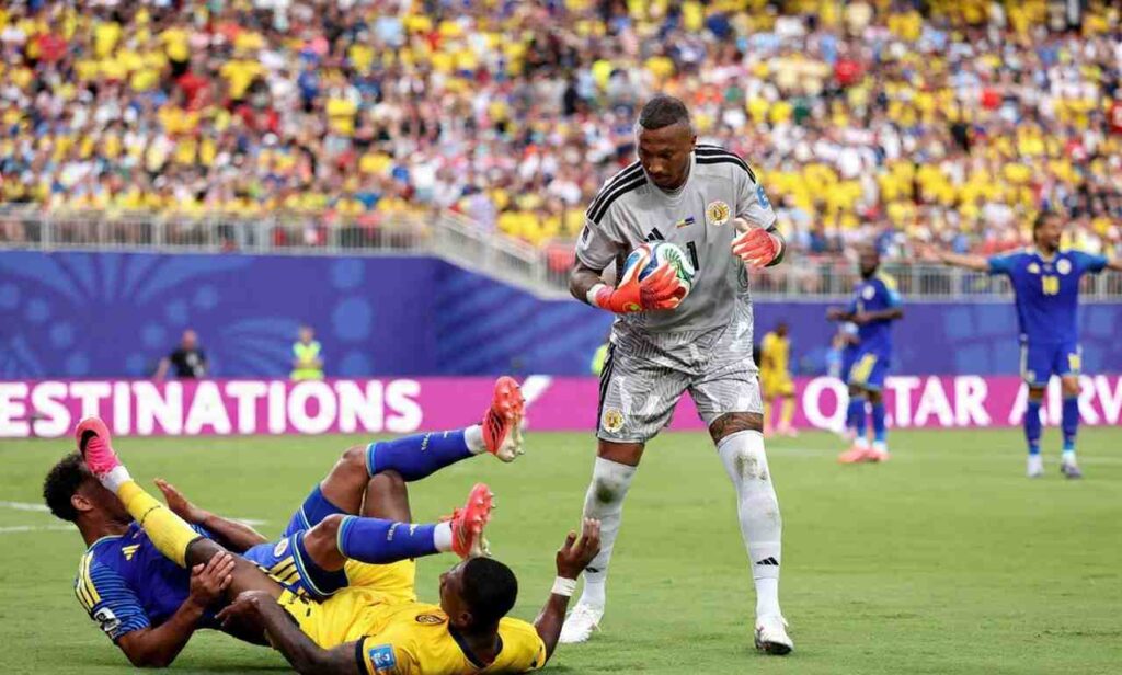 Eloy Room Trybal News Curaçao goalkeeper Eloy Room wearing number 1 gray jersey catches the ball mid air Behind him Curaçao defender Jurien Gaari 3 in blue jersey collides with Ecuadors Pervis Estupinan 7 in yellow jersey on the grass Packed stadium crowd in yellow and blue fills the background at Kansas City Stadium trybalnews