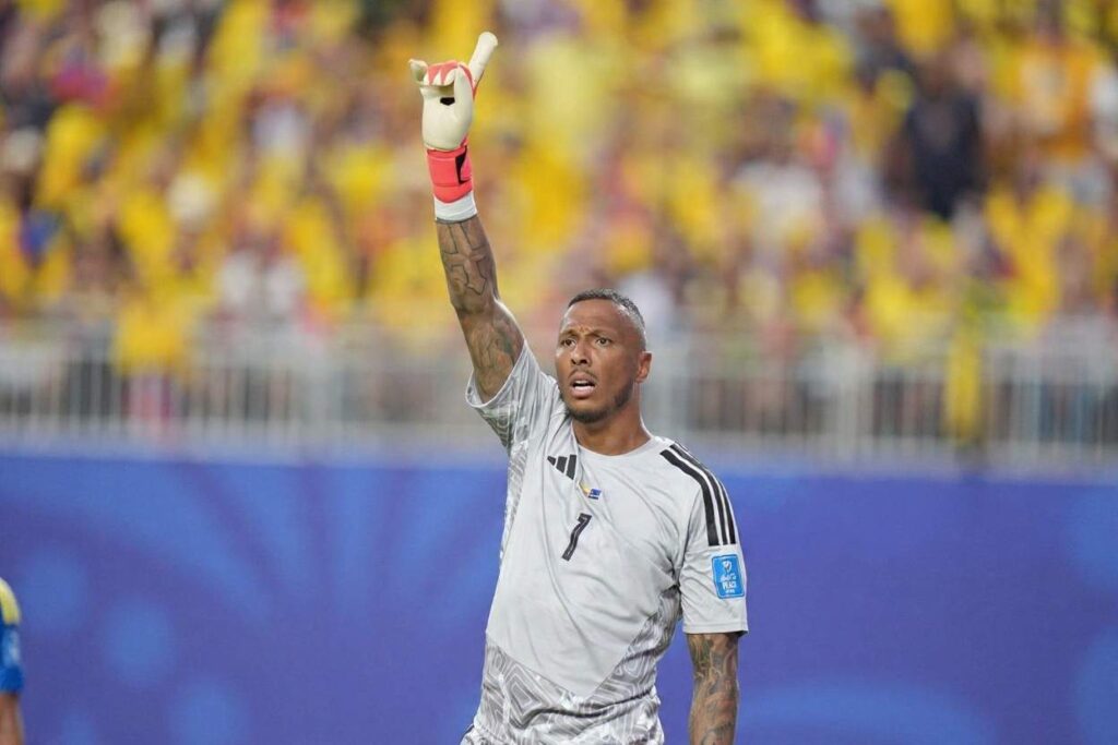 Eloy Room Trybal News Curaçao goalkeeper Eloy Room wearing a light grey jersey with black number 1 reacts with his right arm raised high and index finger pointing upward He has tattoos on both arms and wears red and white goalkeeper gloves His grey kit has black Adidas stripes on the shoulders and the Curaçao Football Federation crest on the chest The background is blurred with a crowd wearing yellow in the stadium stands and blue advertising boards below them Match was FIFA World Cup 2026 Group E Ecuador vs Curaçao at Arrowhead Stadium Kansas City Stadium Kansas City Missouri United States trybalnews