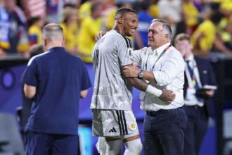 Curaçao goalkeeper Eloy Room, wearing a light grey goalkeeper jersey with number 1 and white shorts, embraces Dick Advocaat, the Curaçao manager and head coach. Room smiles toward the right while Advocaat, wearing a white button-down shirt, dark trousers, and a FIFA World Cup accreditation lanyard, has his left arm around Room’s shoulder and his right hand on Room’s arm. Both appear to be sharing a positive moment at full time. The background is blurred with stadium seats and fans wearing yellow. Match was FIFA World Cup 2026 Group E, Ecuador vs Curaçao at Kansas City Stadium, Kansas City, Missouri, United States on June 20, 2026.