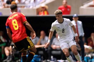 Soccer player in white (number 8) dribbling the ball toward the goal while guarded by a opponent in red jersey on a sunny field.