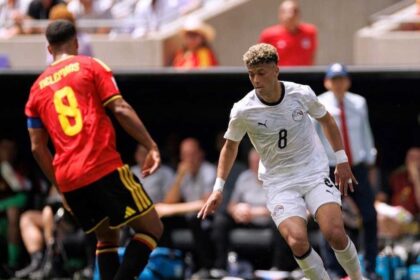 Soccer player in white (number 8) dribbling the ball toward the goal while guarded by a opponent in red jersey on a sunny field.