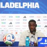 Man at a press conference in front of a blue backdrop reading PHILADELPHIA with a soccer ball microphone and water bottles on the table and a Philadelphia branded laptop nearby  trybalnews