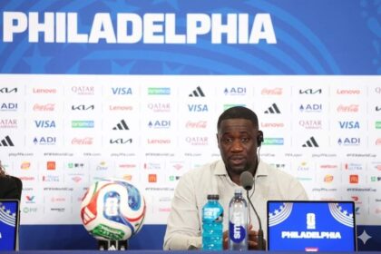 Man at a press conference in front of a blue backdrop reading 'PHILADELPHIA,' with a soccer ball, microphone, and water bottles on the table and a Philadelphia-branded laptop nearby.