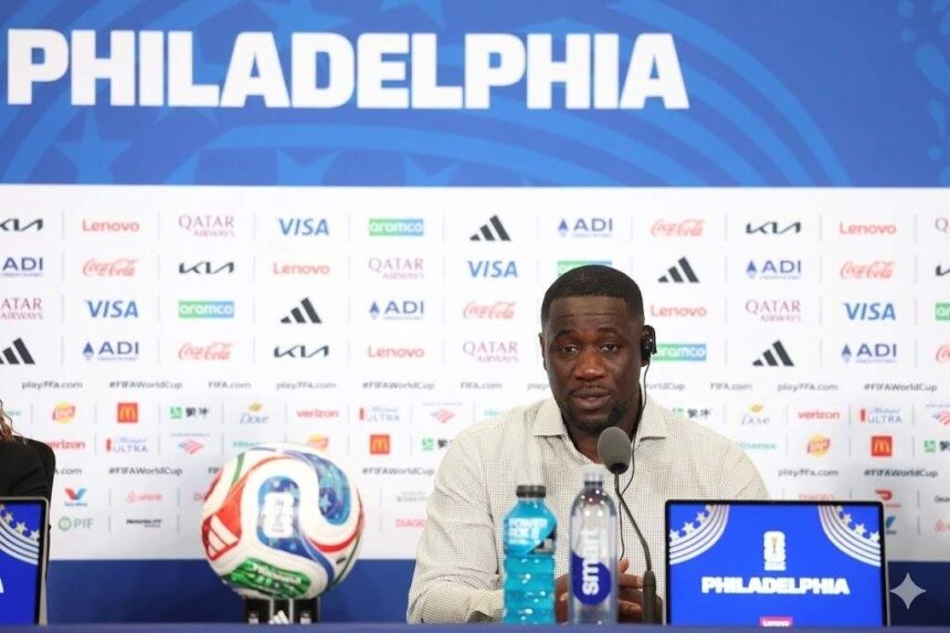 Man at a press conference in front of a blue backdrop reading PHILADELPHIA with a soccer ball microphone and water bottles on the table and a Philadelphia branded laptop nearby  trybalnews