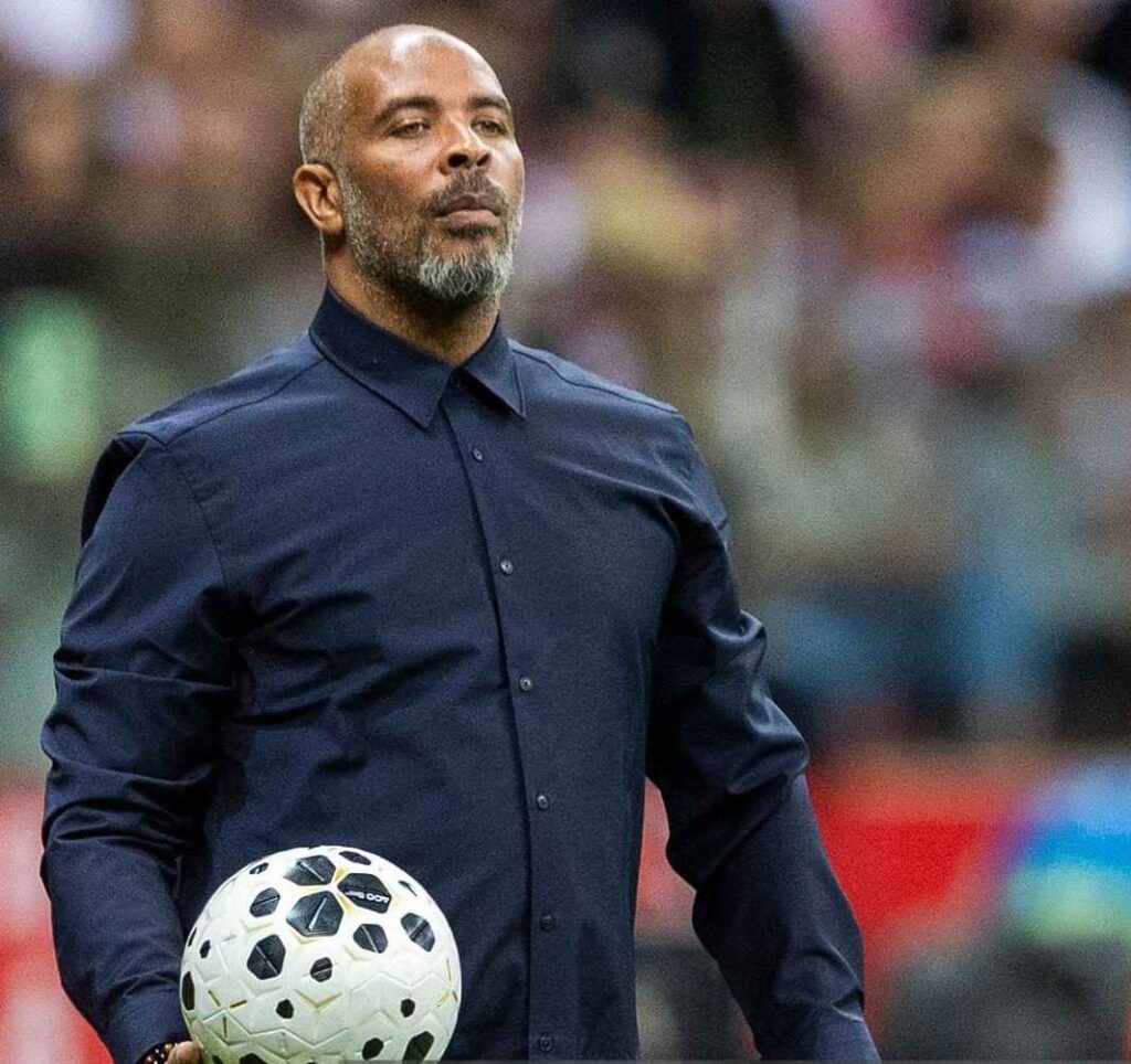 EricChelle Trybal News Eric Chelle in a dark blue button down shirt holding a soccer ball on a field with a blurred crowd in the background trybalnews