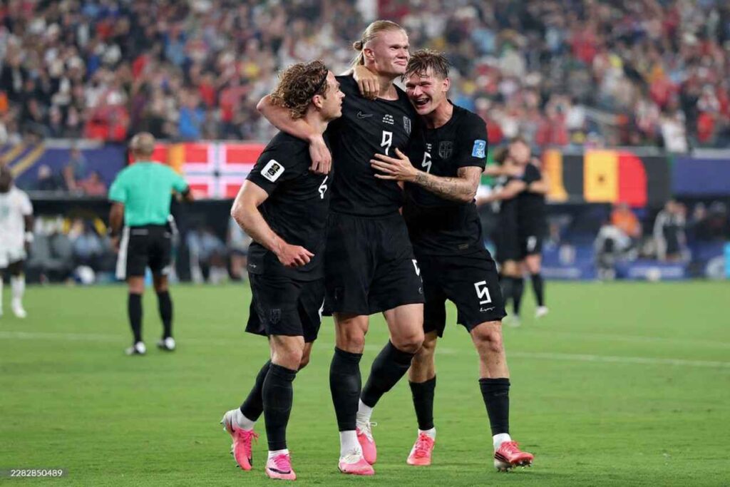 Erling Haaland tall with blond hair in a bun wears Norways all black 9 jersey and shorts He hugs two teammates as they celebrate together on the pitch One teammate has curly brown hair and a headband the other has short brown hair and visible arm tattoos All wear pink football boots The match is Norway vs Senegal at New York New Jersey Stadium FIFA World Cup 2026 Group I June 22 2026 East Rutherford New Jersey A packed stadium crowd and Norwegian flag are blurred in the background  trybalnews