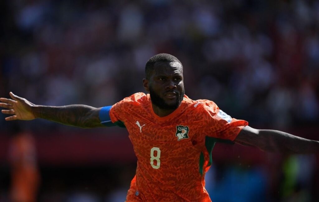 FrankKessie Trybal News Ivory Coast midfielder Franck Kessié wearing orange jersey number 8 with a blue captains armband on his left arm celebrates after scoring He stands with both arms stretched wide to his sides looking focused toward the right side of the frame His orange kit has a leopard pattern and the Ivory Coast crest on the chest The background is blurred with stadium seats and spectators in red and blue Match was FIFA World Cup 2026 Group E Germany vs Ivory Coast at BMO Field Toronto Stadium Toronto Ontario Canada on June 20 2026 trybalnews