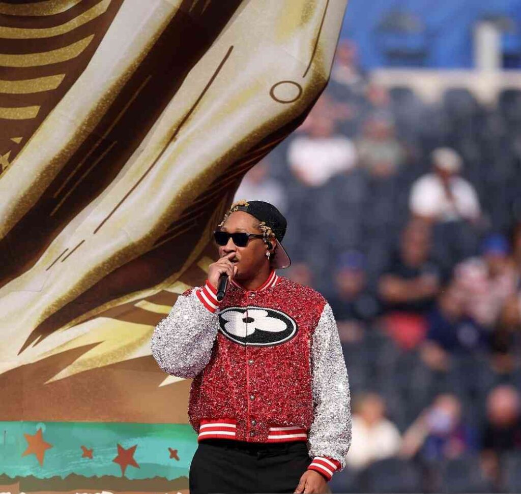 Future Trybal News Male performer Future wearing a red sequined jacket with white sleeves sunglasses and a backward cap singing into a microphone on a stadium stage with a large World Cup sculpture behind him at the 2026 FIFA World Cup Opening Ceremony trybalnews