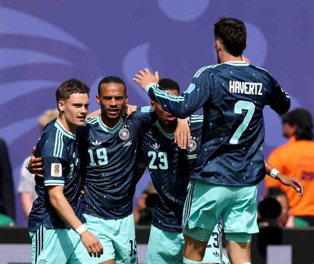 Germany Players Trybal News Germany forward Leroy Sane wearing navy 19 jersey with light blue trim and light blue shorts celebrates with teammates after scoring Sane is in the center with his arm around a teammate looking forward with a focused expression To his right Kai Havertz wearing navy 7 jersey with HAVERTZ on the back puts his hand on Saneaposs shoulder To Saneaposs left a teammate in 21 and another in 23 embrace the group All wear matching navy Adidas kits with Germany crest and light blue shorts FIFA World Cup 2026 Group E match between Ecuador and Germany at New York New Jersey Stadium East Rutherford New Jersey on June 25 2026 trybalnews