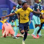 Ecuador forward Gonzalo Plata wearing yellow 19 jersey with navy trim matching navy shorts and navy socks celebrates after scoring He has short curly black hair mouth open in celebration and points with both arms extended to the sides while running on the green pitch Tattoos are visible on his right arm and he wears a white wristband on his left wrist FIFA World Cup 2026 Group E match between Ecuador and Germany at New York New Jersey Stadium East Rutherford New Jersey on June 25 2026 Ecuador crest and Marathon sponsor logo visible on kit Germany goalkeeper in orange kit is on the ground behind him  trybalnews
