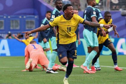 Ecuador forward Gonzalo Plata wearing yellow #19 jersey with navy trim, matching navy shorts, and navy socks celebrates after scoring. He has short curly black hair, mouth open in celebration, and points with both arms extended to the sides while running on the green pitch. Tattoos are visible on his right arm and he wears a white wristband on his left wrist. FIFA World Cup 2026 Group E match between Ecuador and Germany at New York New Jersey Stadium, East Rutherford, New Jersey on June 25, 2026. Ecuador crest and Marathon sponsor logo visible on kit. Germany goalkeeper in orange kit is on the ground behind him.