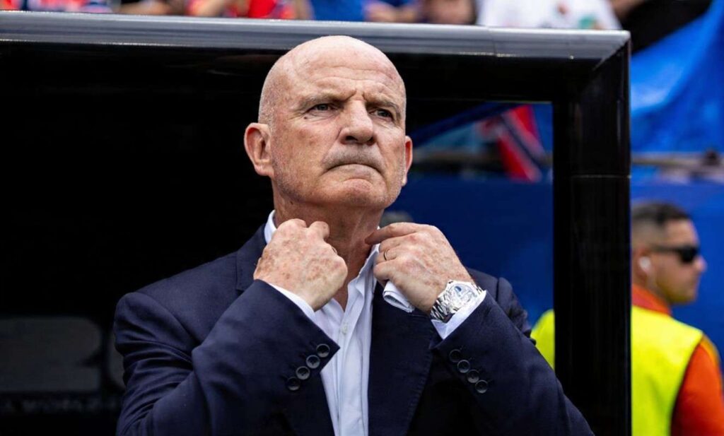 Guy Stephan Trybal News Didier Deschamps and France assistant coach Guy Stephan a bald older man with a serious expression stands in the dugout adjusting the collar of his white shirt with both hands He wears a dark navy suit jacket and a large silver wristwatch on his left wrist 2026 FIFA World Cup Group I match between Norway and France on June 26 2026 at Boston Stadium in Foxborough Massachusetts A black dugout frame blurred blue stadium seating and a staff member in a yellow vest are visible in the background trybalnews