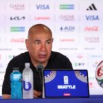 Man at a sports press conference with a Seattle laptop water bottles and a soccer ball on the table behind him  trybalnews