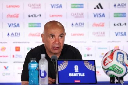 Man at a sports press conference with a Seattle laptop, water bottles, and a soccer ball on the table behind him.