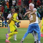 South Africa head coach Hugo Broos with white hair wearing white shirt and blue patterned shorts hugs a Bafana Bafana player wearing a brown FIFA World Cup 2026 training bib over yellow kit Broos smiles broadly while embracing the player Other South African players in yellow kits with green trim and brown training bibs celebrate around them on the pitch including one player jumping with arms around a staff member FIFA World Cup 2026 scene after Bafana Bafana progressed to knockout round for first time ever  trybalnews