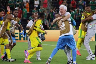 South Africa head coach Hugo Broos with white hair, wearing white shirt and blue patterned shorts, hugs a Bafana Bafana player wearing a brown FIFA World Cup 2026 training bib over yellow kit. Broos smiles broadly while embracing the player. Other South African players in yellow kits with green trim and brown training bibs celebrate around them on the pitch, including one player jumping with arms around a staff member. FIFA World Cup 2026 scene after Bafana Bafana progressed to knockout round for first time ever.