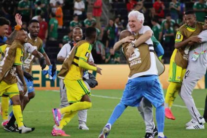 South Africa head coach Hugo Broos with white hair, wearing white shirt and blue patterned shorts, hugs a Bafana Bafana player wearing a brown FIFA World Cup 2026 training bib over yellow kit. Broos smiles broadly while embracing the player. Other South African players in yellow kits with green trim and brown training bibs celebrate around them on the pitch, including one player jumping with arms around a staff member. FIFA World Cup 2026 scene after Bafana Bafana progressed to knockout round for first time ever.
