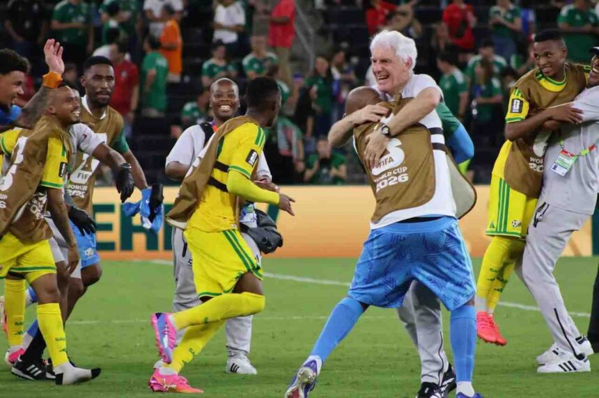 South Africa head coach Hugo Broos with white hair wearing white shirt and blue patterned shorts hugs a Bafana Bafana player wearing a brown FIFA World Cup 2026 training bib over yellow kit Broos smiles broadly while embracing the player Other South African players in yellow kits with green trim and brown training bibs celebrate around them on the pitch including one player jumping with arms around a staff member FIFA World Cup 2026 scene after Bafana Bafana progressed to knockout round for first time ever  trybalnews
