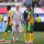 South Africa head coach Hugo Broos an older man with white hair wearing a grey Adidas tracksuit walks on the pitch consoling players after elimination South Africa players in yellow jerseys with green shorts and yellow socks stand nearby including two players embracing with one wearing a purple training bib Another player in yellow and green walks away on the left The 2026 FIFA World Cup Round of 32 match between South Africa and Canada was played on June 28 2026 at Los Angeles Stadium SoFi Stadium in Inglewood California A goal net and stadium advertising boards are visible in the background  trybalnews