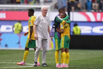 South Africa head coach Hugo Broos, an older man with white hair wearing a grey Adidas tracksuit, walks on the pitch consoling players after elimination. South Africa players in yellow jerseys with green shorts and yellow socks stand nearby, including two players embracing with one wearing a purple training bib. Another player in yellow and green walks away on the left. The 2026 FIFA World Cup Round of 32 match between South Africa and Canada was played on June 28, 2026 at Los Angeles Stadium, SoFi Stadium, in Inglewood, California. A goal net and stadium advertising boards are visible in the background.