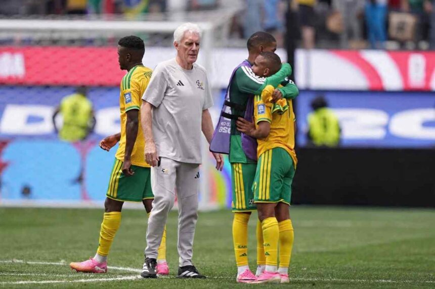 South Africa head coach Hugo Broos an older man with white hair wearing a grey Adidas tracksuit walks on the pitch consoling players after elimination South Africa players in yellow jerseys with green shorts and yellow socks stand nearby including two players embracing with one wearing a purple training bib Another player in yellow and green walks away on the left The 2026 FIFA World Cup Round of 32 match between South Africa and Canada was played on June 28 2026 at Los Angeles Stadium SoFi Stadium in Inglewood California A goal net and stadium advertising boards are visible in the background  trybalnews