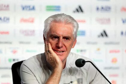 Hugo Broos, white-haired South Africa head coach in a light gray shirt, sits at a press conference table with a microphone. He rests his chin on his hand with a thoughtful expression. Sponsor logos are blurred in the background. Atlanta Stadium, FIFA World Cup 2026 Group A, June 18 2026.