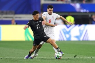 Two soccer players compete for the ball during a match, one in black kit (number 11) shielding the ball from a white-kit opponent (number 6).