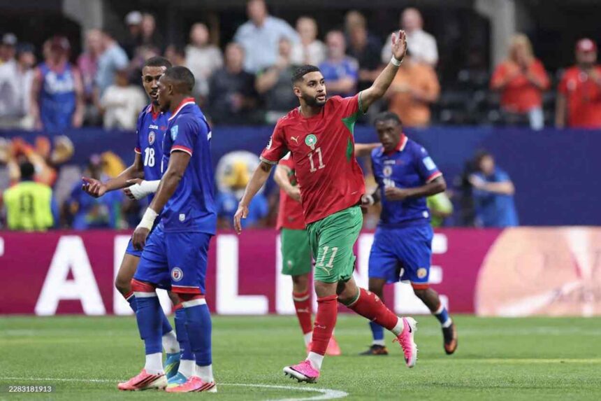 Morocco midfielder Ismael Saibari wearing red 11 jersey with green trim and green shorts celebrates with right arm raised He has a beard short hair white wristband and pink Nike boots with red socks Haiti players in blue kits with 18 visible stand nearby on the pitch Blurred maroon advertising board with white text and stadium crowd in background during FIFA World Cup 2026 Group C match between Morocco and Haiti at Atlanta Stadium Atlanta Georgia on June 24 2026  trybalnews