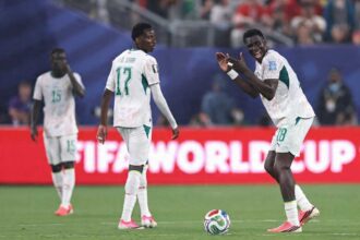 Ismaila Sarr in Senegal’s white #18 jersey with green trim claps both hands together and shouts in celebration on the pitch. A football sits on the grass near his pink and black boots. Behind him, P.M. Saar #17 in a white long-sleeve jersey looks over his shoulder, and teammate #15 covers his mouth with his hand. The match is Norway vs Senegal at New York New Jersey Stadium, FIFA World Cup 2026 Group I, June 22 2026, East Rutherford New Jersey. A red "FIFA WORLD CUP" advertising board is visible behind them.