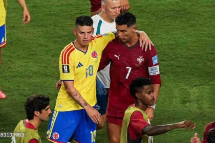 Colombia midfielder James Rodriguez, wearing a yellow kit with number 10 and blue shorts, stands next to Portugal forward Cristiano Ronaldo, wearing a red kit with number 7. Rodriguez has his arm around Ronaldo’s shoulder in a post-match moment. A staff in white and other players in training bibs are visible on the green pitch. The 2026 FIFA World Cup Group K match between Colombia and Portugal took place on June 27, 2026 at Miami Stadium in Miami, United States.