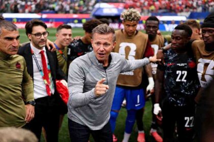 Canada head coach Jesse Marsch, wearing a grey quarter-zip with the Canada Soccer crest, speaks and points to his players in a post-match huddle. Staff and players surround him, including a man in a green Canada top on the left, a staff member in a suit with a red tie, and Canada players in black speckled kits and brown training bibs, with #22 visible. The 2026 FIFA World Cup Round of 32 match between South Africa and Canada was played on June 28, 2026 at Los Angeles Stadium in Inglewood, California, with Canada advancing to the Round of 16. A packed stadium crowd is blurred in the background.