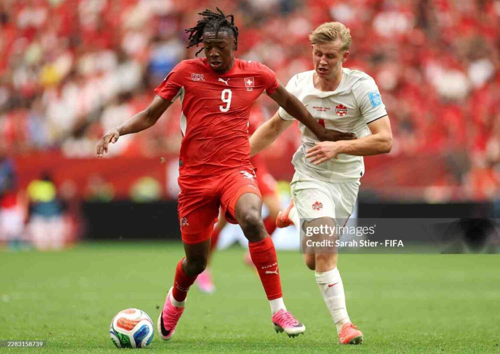 JohanManzambi Trybal News Switzerland forward Johan Manzambi wearing red 9 jersey with dreadlocks controls the World Cup match ball while Canada defender Luc de Fougerolles in white 4 jersey challenges with his hand on Manzambiaposs shoulder Both players wear pinkorange boots on green pitch with blurred red crowd in background during FIFA World Cup 2026 Group B match between Switzerland and Canada at BC Place Vancouver on June 24 2026 trybalnews