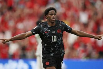 Jonathan David of Canada celebrates after scoring Canada's second goal vs Qatar at FIFA World Cup 2026, BC Place Vancouver, June 18, 2026. He wears a black Canada kit #10 with arms spread wide, crowd blurred red behind him.