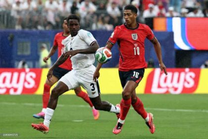 England midfielder Jude Bellingham, wearing a red kit with number 10 and bright pink boots, runs with the ball. He is challenged by Panama defender Jose Cordoba, number 3 in a white kit, who is lunging to tackle. Another England player in red is visible behind them. The 2026 FIFA World Cup Group L match between Panama and England took place on June 27, 2026 at New York/New Jersey Stadium in East Rutherford. Lay’s advertising boards and a packed stadium crowd are in the background.