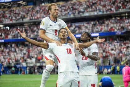 Three England footballers celebrate a goal on the pitch, one lifted on his teammate's shoulders as others raise their arms in jubilation, stadium full of fans behind them in win over Croatia.