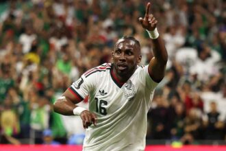 Mexico forward Julian Quinones wearing white #16 jersey with green and red trim, matching white shorts, and white wristbands celebrates after scoring. He has short braided hair, looks upward, and points his right index finger to the sky while running on the green pitch. Left arm is bent at his side. FIFA World Cup 2026 Group A match between Czechia and Mexico at Mexico City Stadium, Mexico City, Mexico on June 24, 2026. Mexico crest and Adidas logo visible on kit with blurred crowd in green behind him.