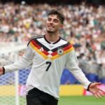 Soccer player in Germany jersey 7 celebrates with arms outstretched on a green field crowd blurred in stadium behind him from a match scene  trybalnews