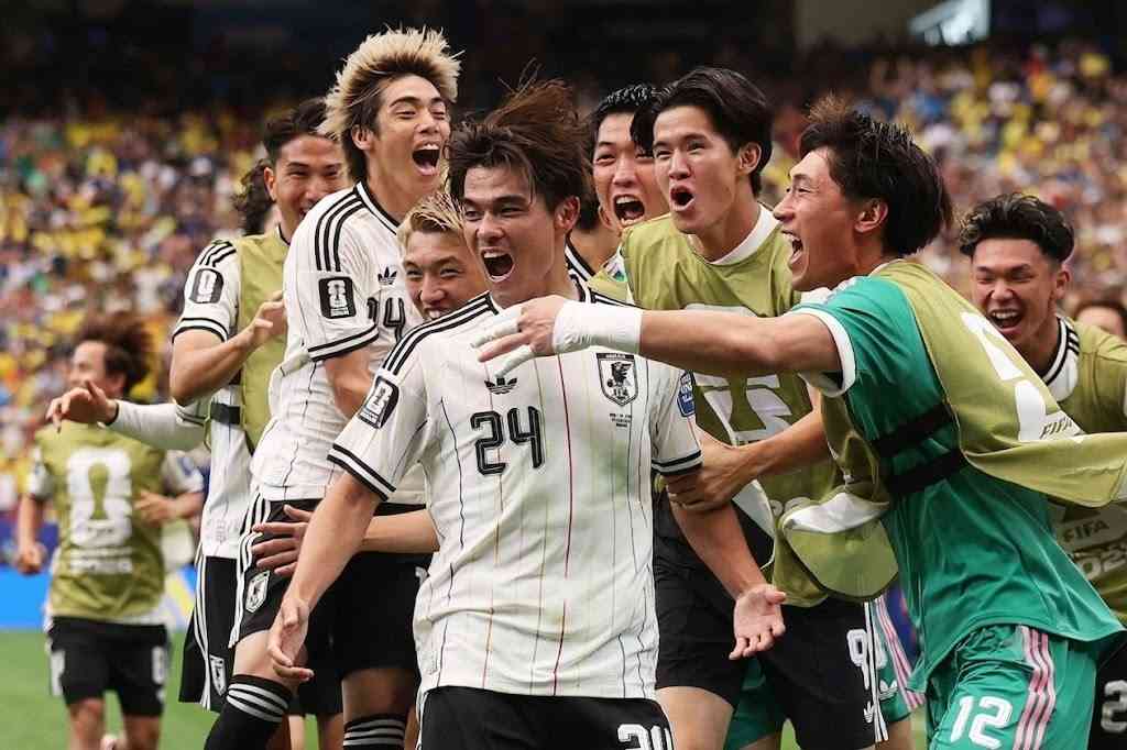 Kaishu Sano Trybal News Japan midfielder Kaishu Sano wearing white 24 with black stripes and the JFA crest celebrates with his teammates after scoring Japans first goal He is surrounded by Japan players in white kits and staff in green training bibs all shouting and embracing on the pitch The FIFA World Cup 2026 Round of 32 match between Brazil and Japan took place on June 29 2026 at Houston Stadium in Houston Texas A packed stadium crowd is blurred in the background trybalnews