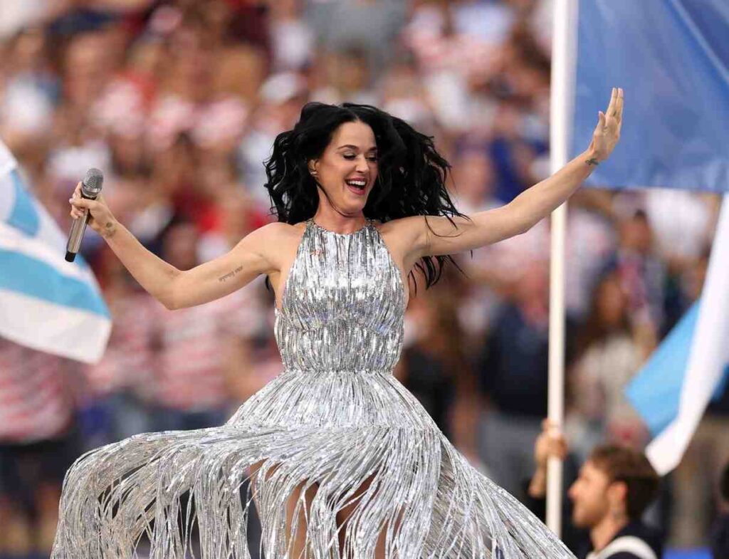 Katy Perry Trybal News Female singer Katy Perry in a silver fringed gown performing on stage with arms raised microphone in hand crowd in the background at 2026 World Cup Opening Ceremony in LA trybalnews