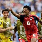 Cabo Verde midfielder Kevin Pina wearing number 6 red jersey with dark sleeves points upward with his right hand and smiles while celebrating Teammates in gold training bibs over blue kits surround him shouting and smiling Stadium crowd blurred in background at Miami Stadium Miami Gardens Florida June 21 2026 during FIFA World Cup 2026 Group H match vs Uruguay  trybalnews