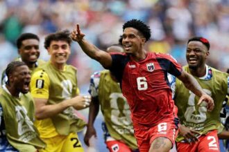 Cabo Verde midfielder Kevin Pina wearing number 6 red jersey with dark sleeves points upward with his right hand and smiles while celebrating. Teammates in gold training bibs over blue kits surround him, shouting and smiling. Stadium crowd blurred in background at Miami Stadium, Miami Gardens, Florida, June 21, 2026 during FIFA World Cup 2026 Group H match vs Uruguay.