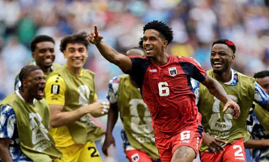 Cabo Verde midfielder Kevin Pina wearing number 6 red jersey with dark sleeves points upward with his right hand and smiles while celebrating Teammates in gold training bibs over blue kits surround him shouting and smiling Stadium crowd blurred in background at Miami Stadium Miami Gardens Florida June 21 2026 during FIFA World Cup 2026 Group H match vs Uruguay  trybalnews