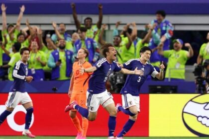 Blue-clad Japanese soccer players celebrate a goal on the field as cheering fans fill the stands behind them.