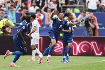 Kylian Mbappé in France’s dark blue #10 jersey runs with both arms raised and index fingers pointing up. He has a captain’s armband on his left arm and wears pink football boots. Teammates and an opponent are visible behind him. The match is France vs Iraq at Philadelphia Stadium during the FIFA World Cup 2026 Group I, June 22 2026, Philadelphia Pennsylvania. Crowd and LED boards are blurred in the background.